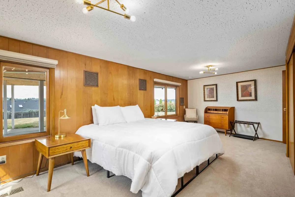 This image shows a serene bedroom with a neatly made bed positioned next to a large window, allowing ample natural light to stream in. A simple dresser and thoughtfully chosen artwork complete the space, adding a sense of personal comfort and calm. The overall palette is soft and neutral, contributing to a restful atmosphere.