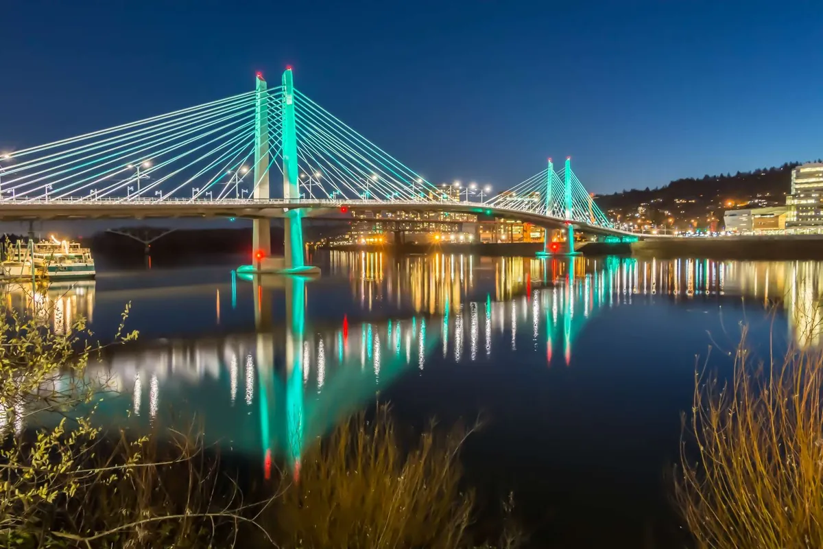 A beautifully illuminated bridge with reflections sparkling on the river below, set against the city skyline at night. The scene is vibrant yet serene, capturing the urban allure of nearby Portland and opportunities for evening outings or photography.