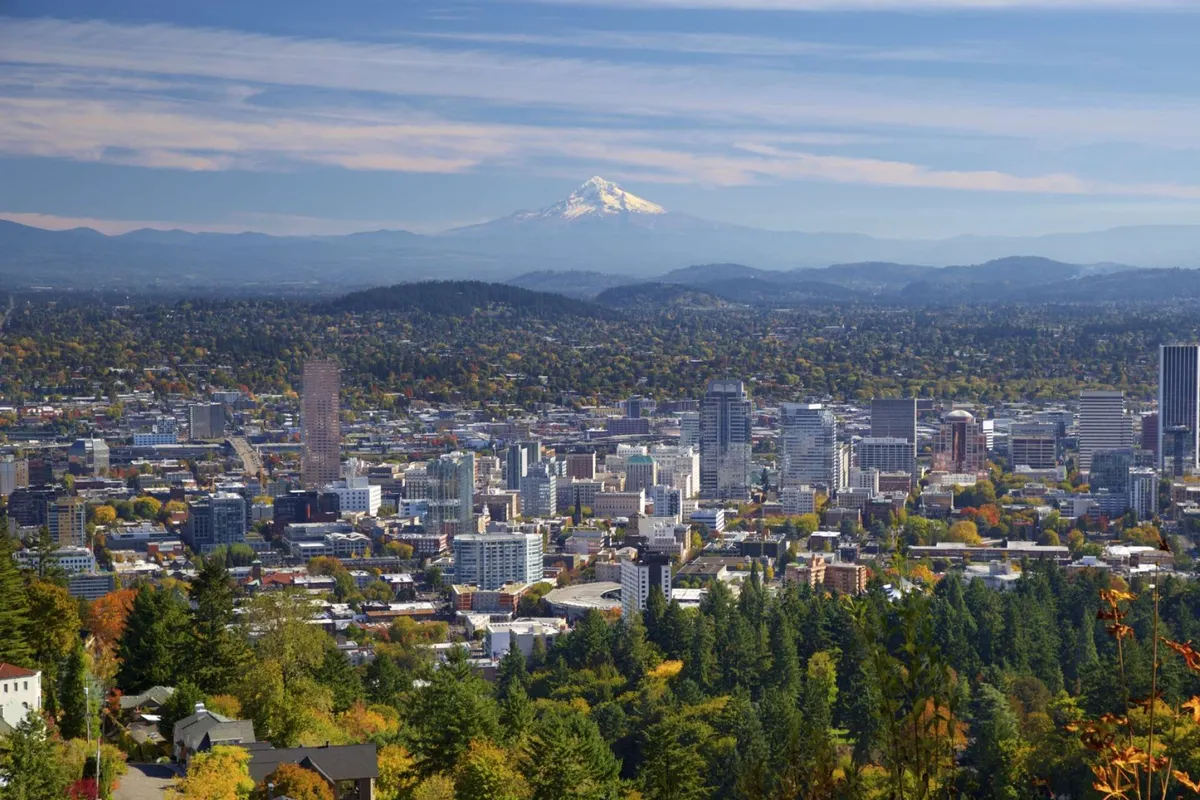 The image features a breathtaking panoramic view of Portland's downtown skyline, Mount Hood in the distance, and vibrant autumn foliage in the foreground. The interplay of cityscape and natural landscape captures the region’s spirit of inspiration and adventure.