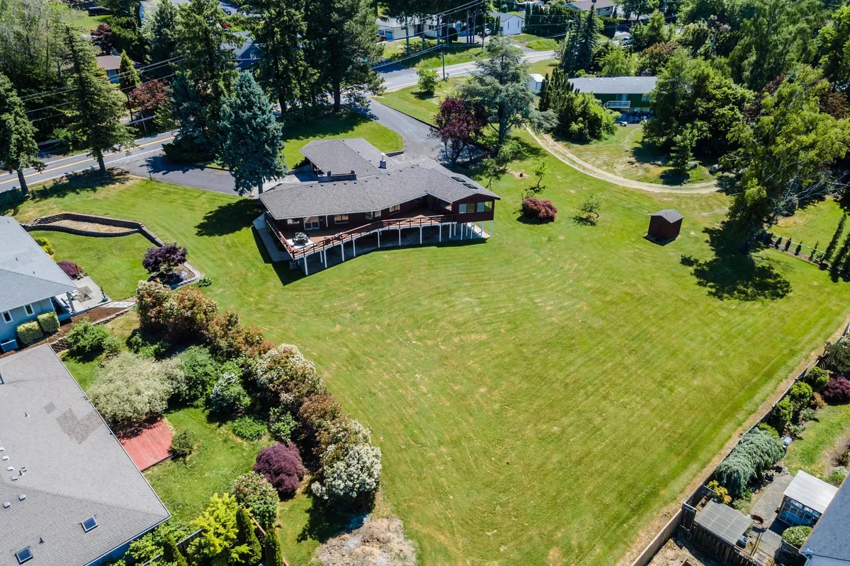 A peaceful, idyllic view of a spacious lawn bordered by a wraparound porch and lush greenery. The neighborhood setting in the background suggests a community-oriented environment, while the large yard offers ample space for pets to roam safely under supervision.