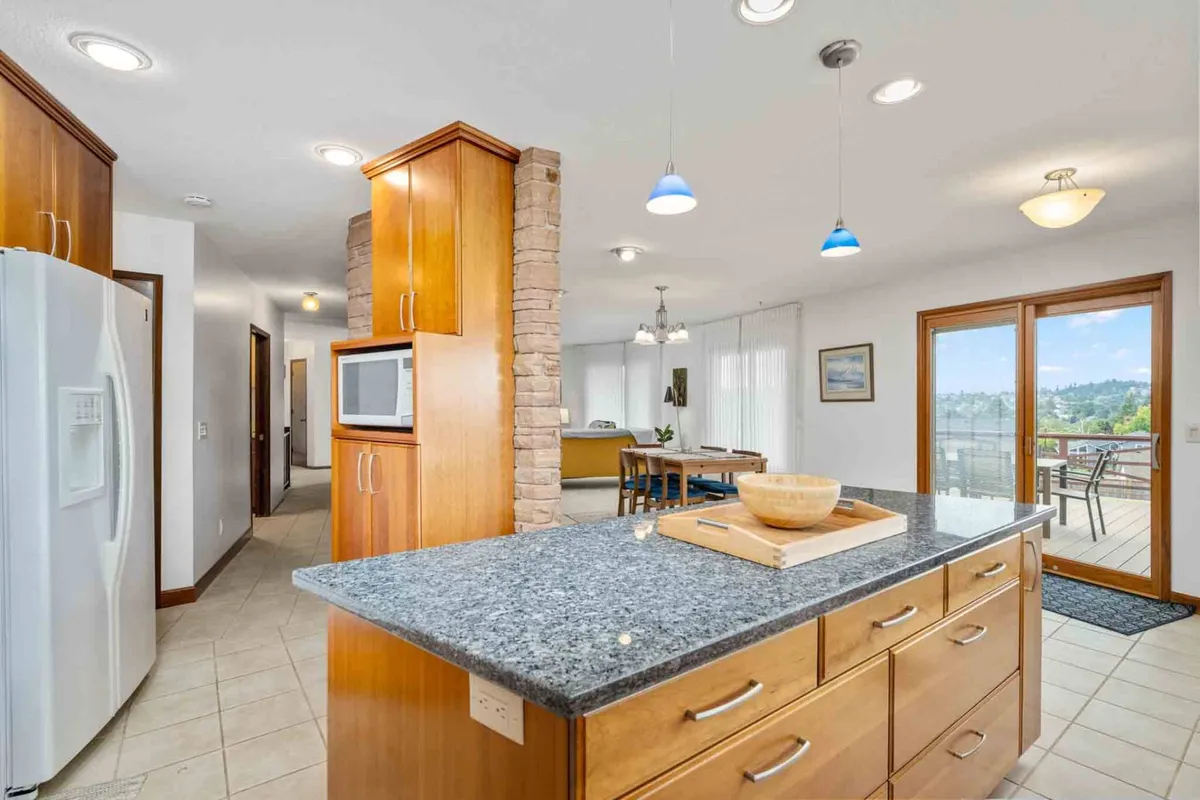 An open kitchen with granite countertops, wooden cabinetry, stainless steel appliances, and a spacious layout that flows into the dining area. The openness encourages both culinary creativity and social interaction, with a glimpse of the outdoors through large windows.