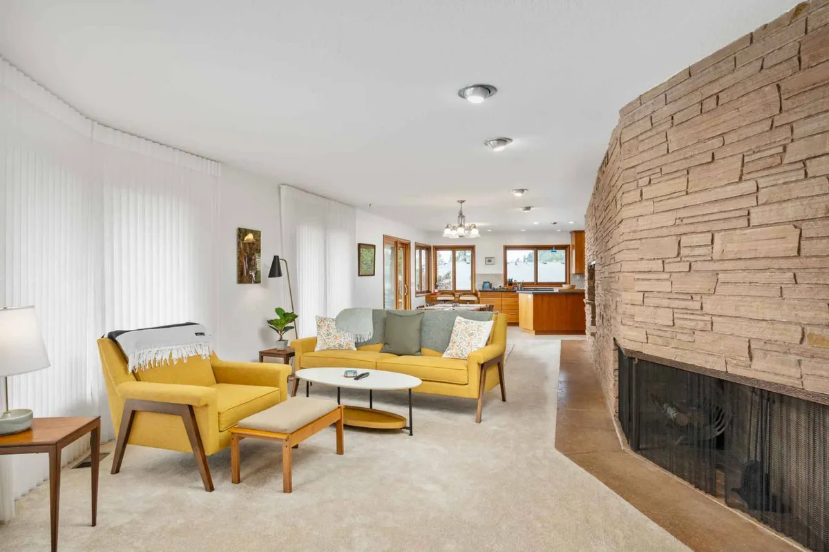 The image showcases a warm, inviting living space with a vibrant yellow sofa, a striking stone fireplace, and a stylish wooden dining area. The layout is open and airy, filled with natural light and touches of mid-century design, creating a perfect setting for relaxing family gatherings or cozy evenings by the fire.