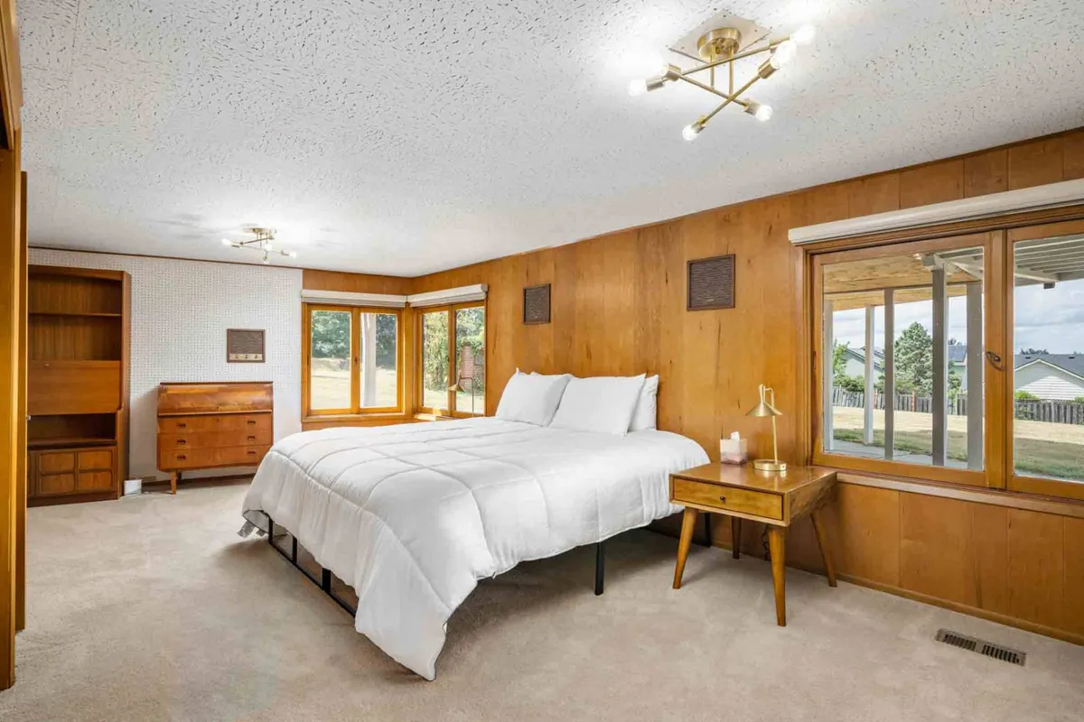 This bedroom features warm wood paneling, a comfy bed positioned beside generous windows, and a functional nightstand. The windows frame relaxing views and let in abundant light, creating an ideal spot for morning reflection or evening stargazing. The setup highlights modern comfort within a tranquil, mid-century inspired design.