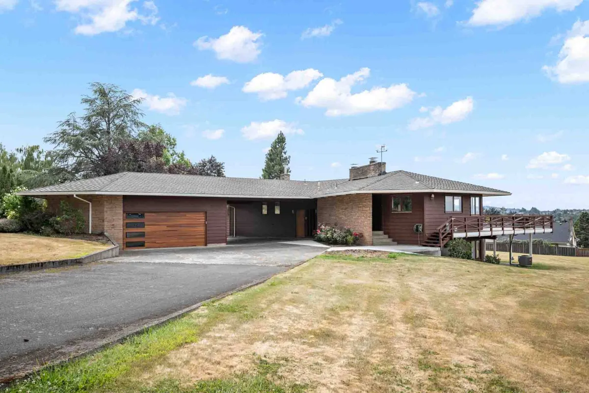 A wide shot of the home's modern exterior, highlighting architectural details, the landscaped yard, a spacious deck with railing, and a welcoming driveway. The environment appears open and inviting, ideal for gatherings or quiet reflection surrounded by nature.