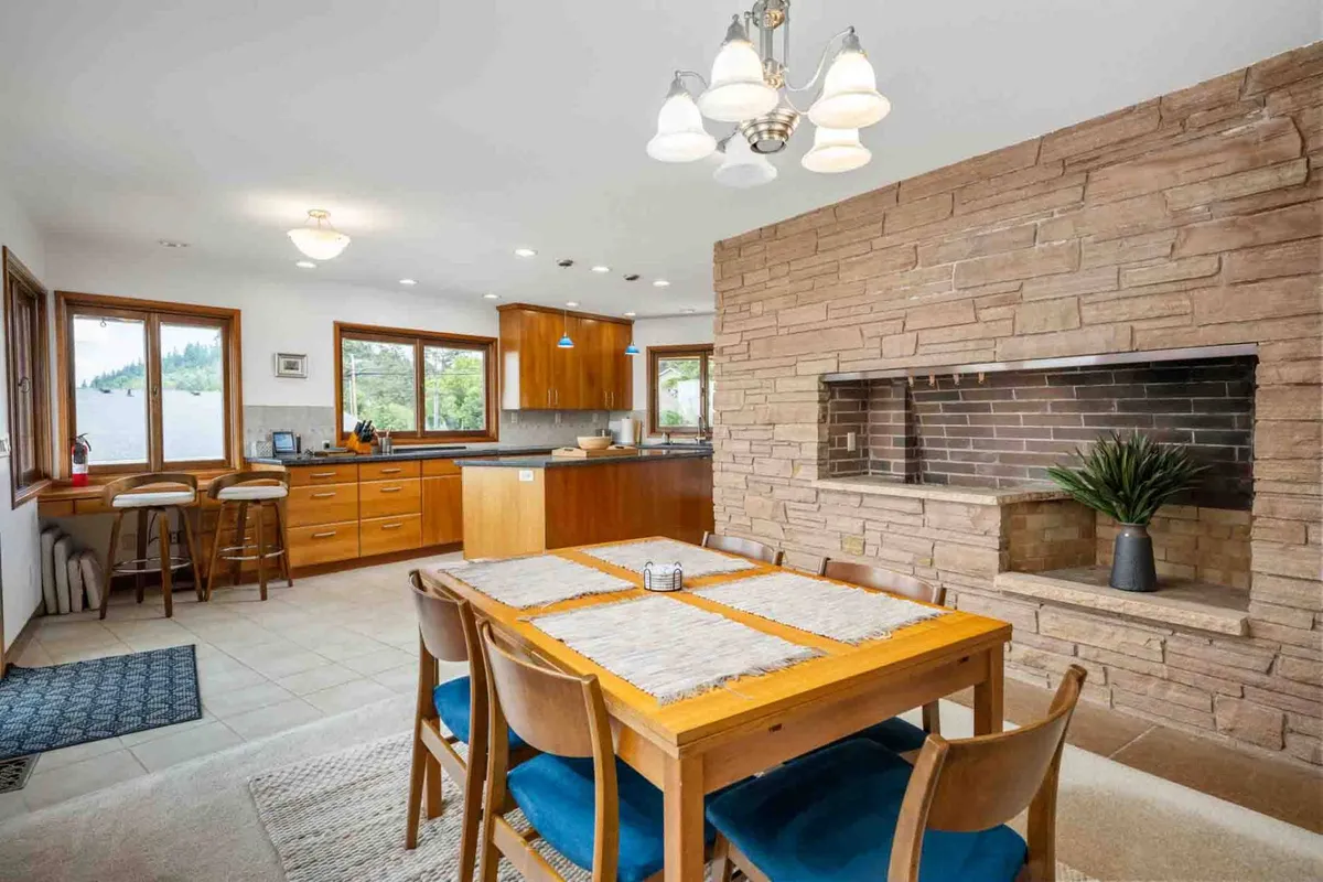 A cozy dining area with a wooden table, adjacent to a kitchen island, set against a stone accent wall and further illuminated by expansive windows. The space is ideal for social gatherings, family meals, and entertaining guests, seamlessly blending elegance with comfort.