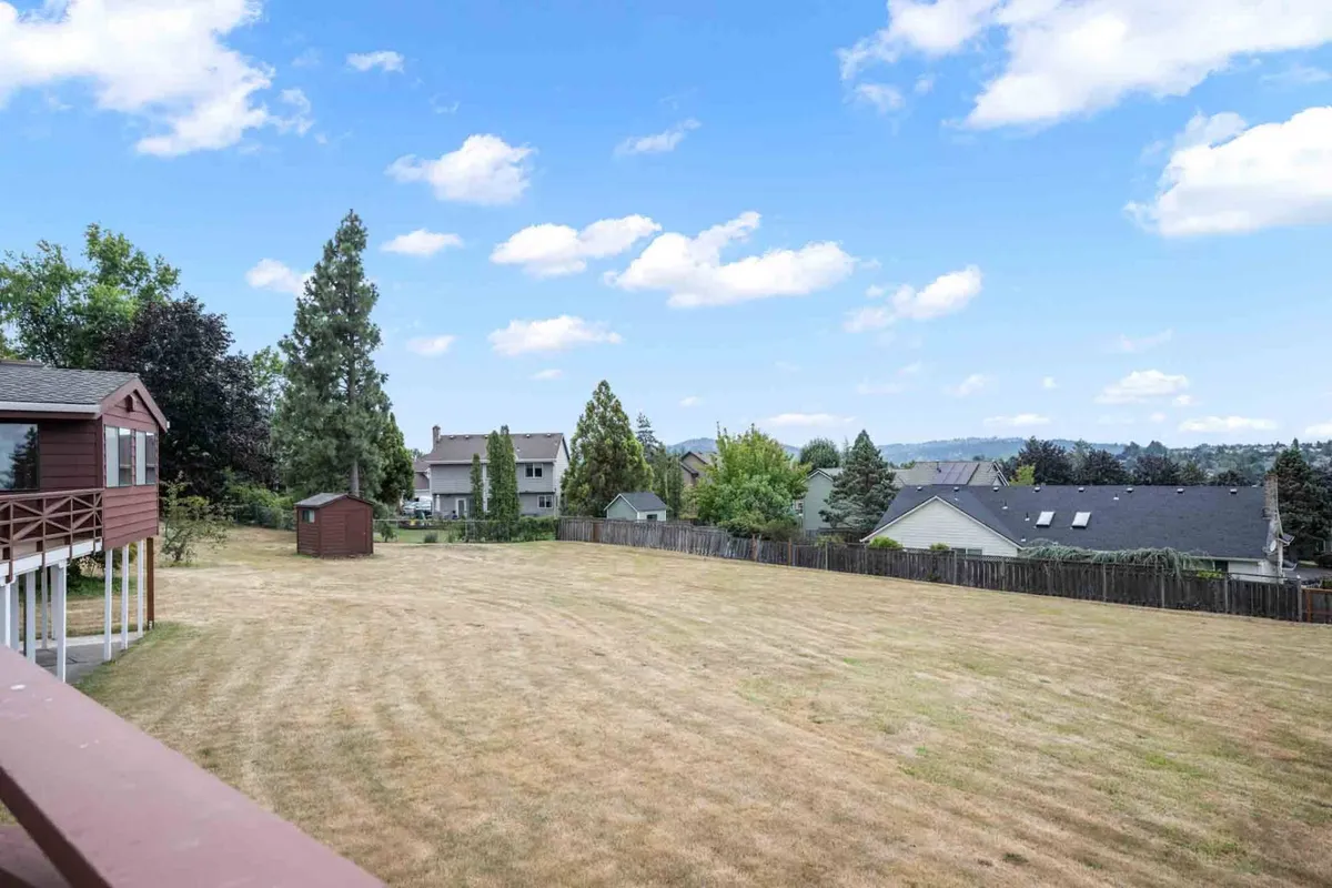 A wide view of a spacious yard bordered by mature trees and distant mountains. Neighboring houses are visible in the background, conveying a sense of community within a backdrop of natural tranquility. The open space and lush greenery highlight the lot's generous size and private feel.