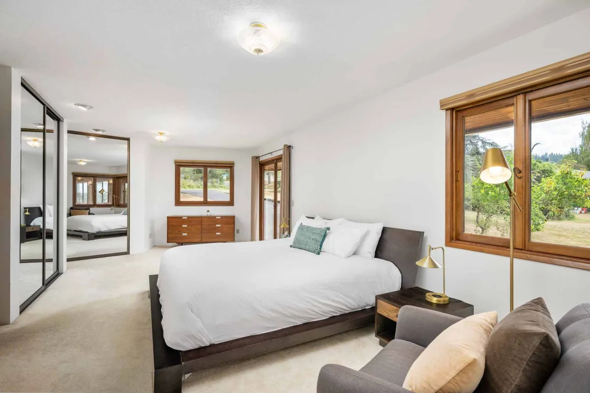 This image presents a cozy bedroom with a soft bed, armchair for reading, and a dresser, all bathed in natural light from a spacious window with a scenic view. The neutral tones and inviting textures create a nurturing environment ideal for relaxation and unwinding after a vibrant day.