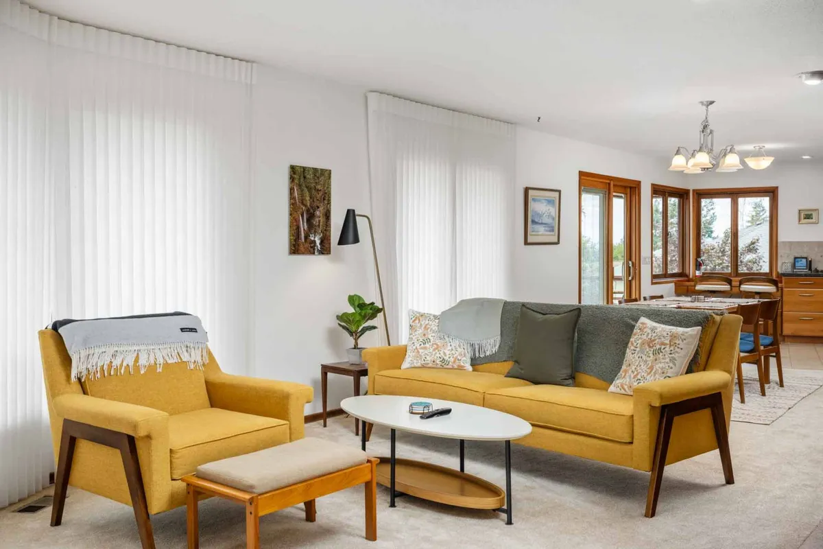 A welcoming living area featuring a vibrant yellow sofa, accent chairs, a modern coffee table, and large panoramic windows that bathe the space in natural light. Decorative artwork and an open floor plan create an inviting, stylish, and sociable environment.