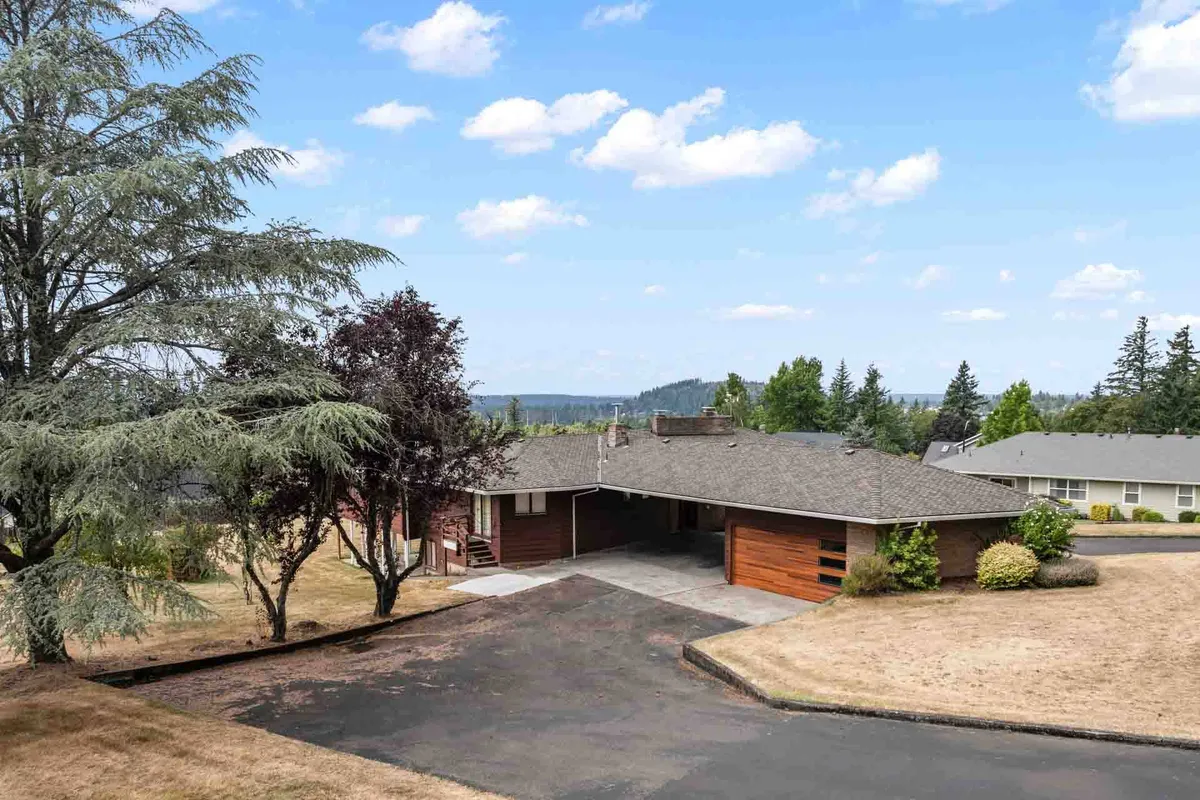 The image showcases the home's striking mid-century modern architecture set against lush greenery. The driveway leads up to the residence, emphasizing its unique roofline and the harmony of the house with its natural surroundings. The landscaping and crisp lines of the structure reflect classic modernist design principles.